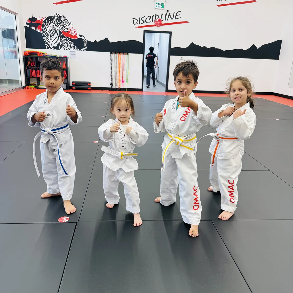 Four young OMAC students in white uniforms posing with fists up on the mats